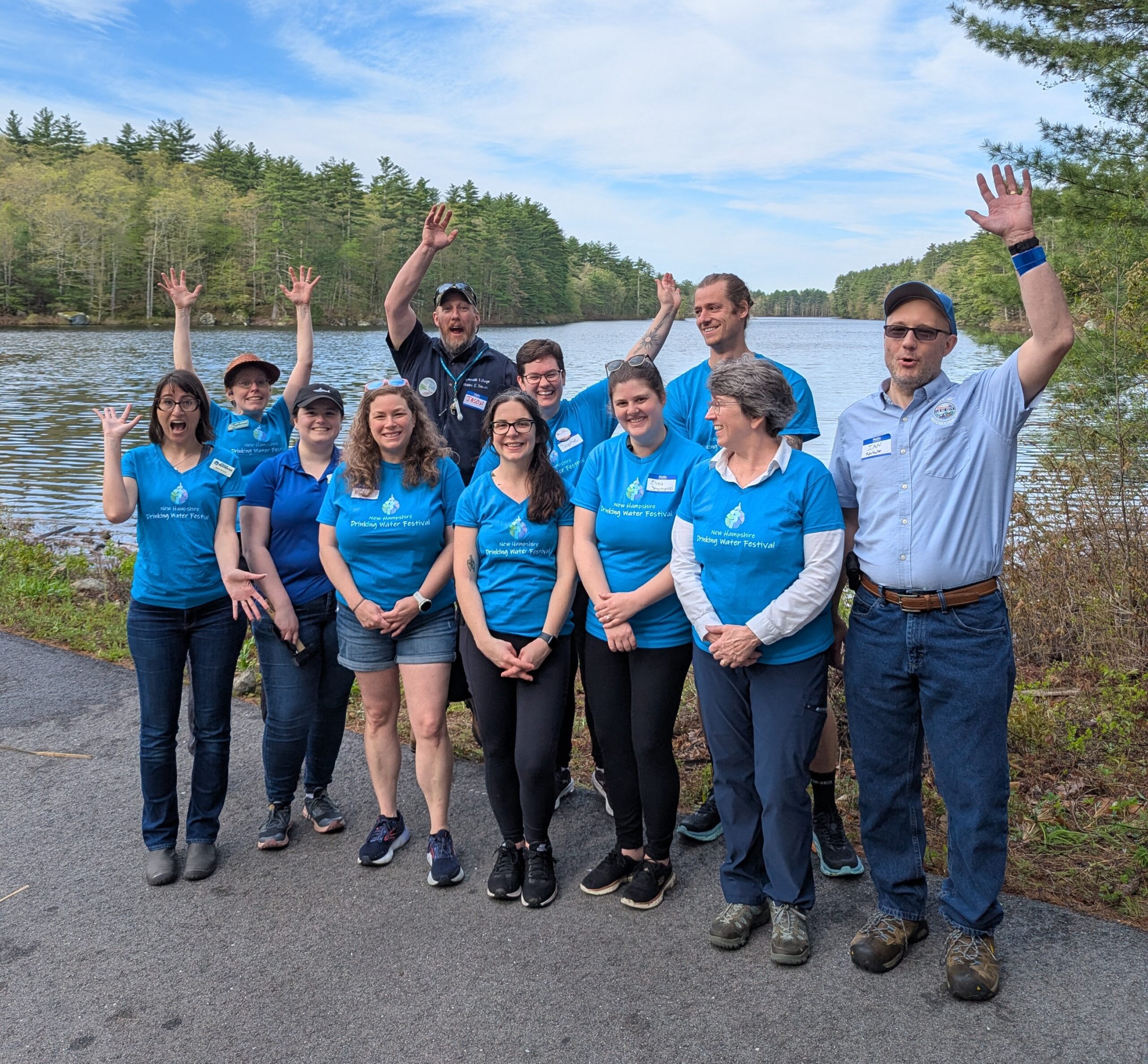 Eleven happy and waving adults in blue shirts standing by a pond.
